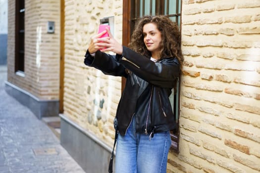 A fashionable and stylish young woman with beautiful curly hair cheerfully poses for a vivid and engaging selfie against a textured urban wall in an exciting environment, capturing a moment of joy