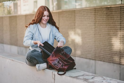 A cheerful young woman sits on a ledge in a vibrant outdoor setting, unpacking her trendy backpack, showcasing her casual style and exuding a relaxed vibe as she enjoys the moment