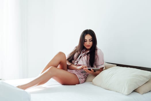 Beautiful brunette woman in pajamas with iPad in white bedroom on bed sleep