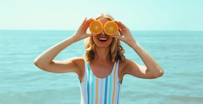 Summer vacation, portrait of happy smiling young woman, laughing, holding oranges fruits, having fun, wearing a swimsuit on the beach on sea coast background