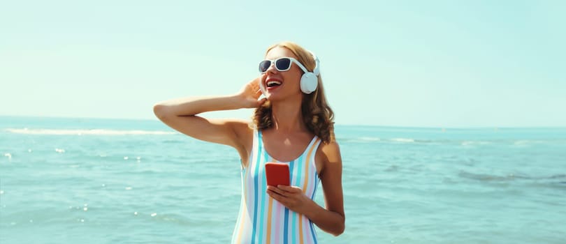 Summer vacation of happy young woman smiles enjoying listening to music with headphones, holding phone, relaxed cheerful girl wear a sunglasses on beach at sea coast with blue sky on a sunny day