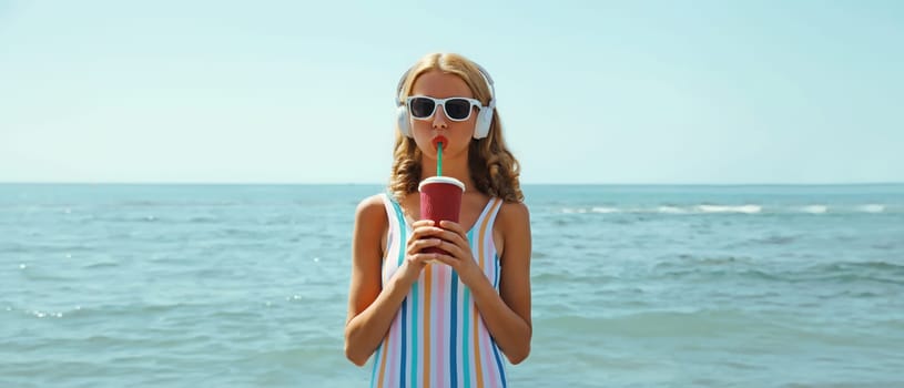 Summer vacation of happy relaxed young woman with cup of fresh juice drink enjoying listening to music with headphones, modern girl wearing a sunglasses on the beach at sea coast background