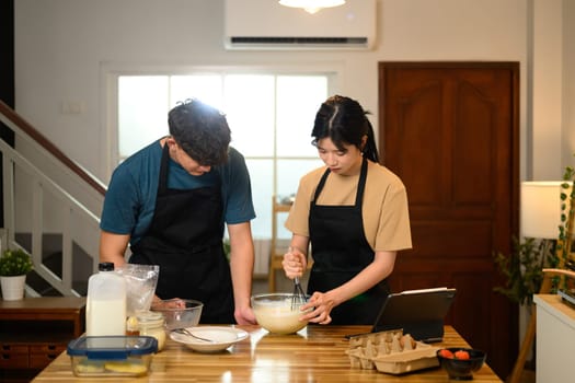 Happy couple in aprons following a recipe while making pancakes at home.