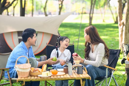 Parents and joyful daughter having a relaxing outdoor meal during family camping trip in nature.