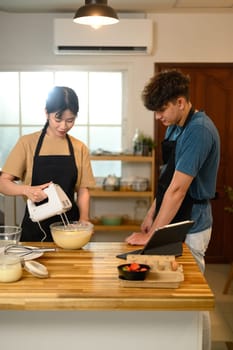 Happy couple in aprons following a recipe while making pancakes at home.