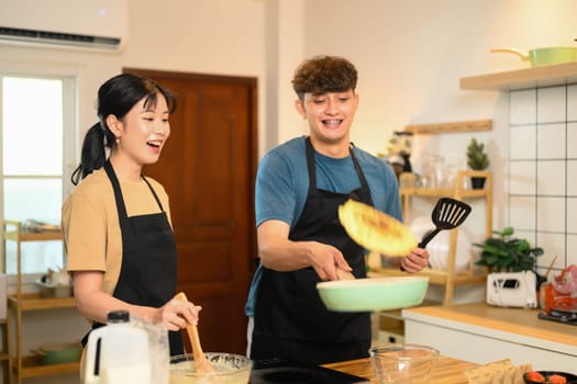 Man flipping pancake in a skillet while his partner watches and laughs.