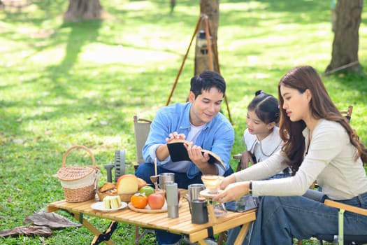 Happy family enjoying a camping trip sitting by the tent with food and browsing on a tablet.