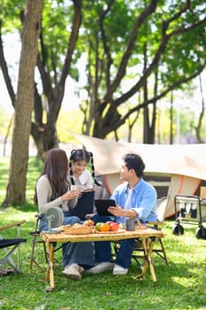 Happy family enjoying a camping trip sitting by the tent with food and browsing on a tablet.