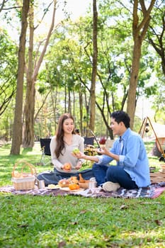 Couple sharing fruit and laughter while camping in a sunny woodland.