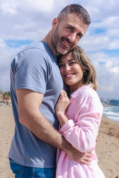 Vertical Portrait of a smiling caucasian young couple embracing on a beach during vacation. Relaxed and affectionate moment by the sea, with blue sky, ocean waves, and sandy shore behind them.