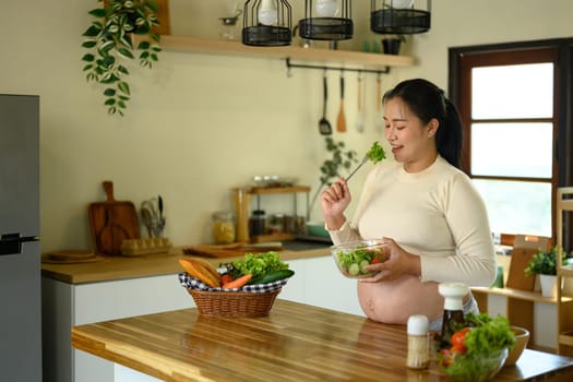 Smiling pregnant woman enjoying fresh vegetable salad in the kitchen. Healthy lifestyle concept.