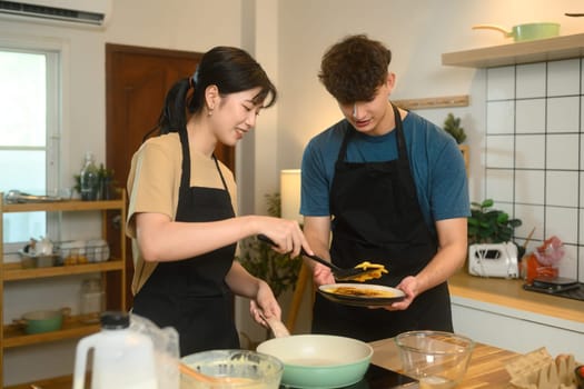 Cheerful young couple preparing breakfast pancakes in cozy home kitchen.
