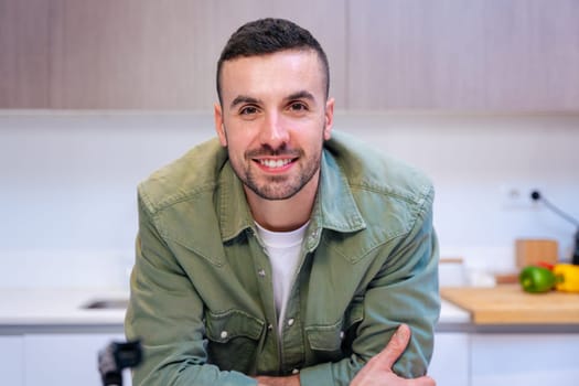 Portrait of a confident young caucasian man smiling at the camera in a stylish and bright kitchen interior, suggesting a relaxed and welcoming home atmosphere with visible food and cooking elements