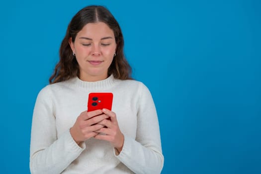 Portrait of a young woman in white sweater looking down at a red smartphone while standing against a vibrant blue background, representing focus,communication,and casual digital lifestyle..Copy space