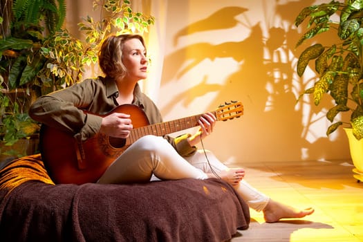 A woman in casual clothes strums an acoustic guitar, relaxing on a cushy surface in a warm, inviting room.