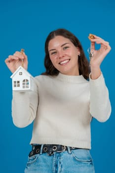 Happy young woman holding a house model and keys, expressing excitement. She wears a white sweater and stands against a blue background. Concept of home ownership,real estate,or investment. Copy space