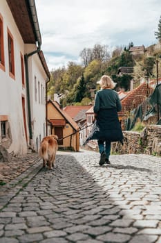 A person strolls through the picturesque streets of Cesky Krumlov alongside a dog, enjoying the beauty of the cobblestone path and surrounding historic architecture under a partly cloudy sky.
