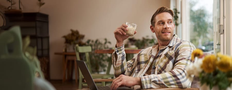Portrait of young freelancer, a man in casual clothes, sitting in cafe and drinking coffee, working remotely on laptop, digital nomad doing his project outdoors in co-working space.