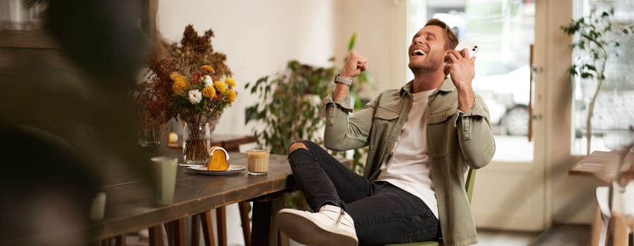 Portrait of relieved, happy young man, listens to happy news or podcast in earphones, sits in coffee shop, holds smartphone, raises hands up and smiling from excitement, spending time in cafe.