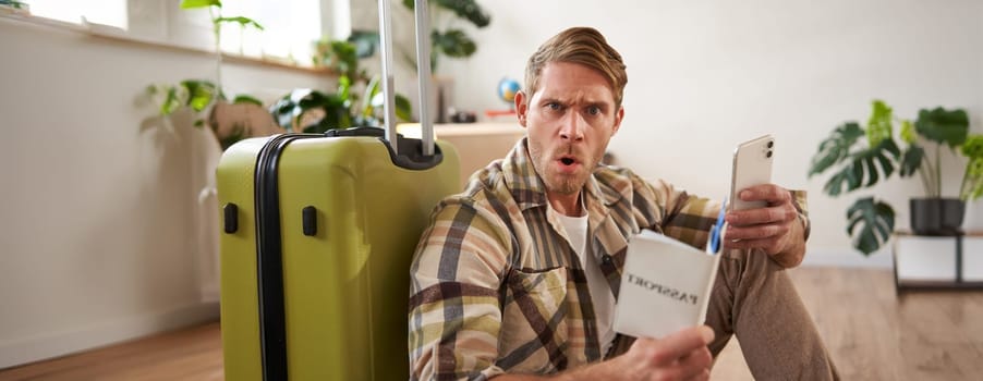 Man looks very confused, using smartphone, holding passport, sitting near luggage, planning his journey or holiday. Copy space