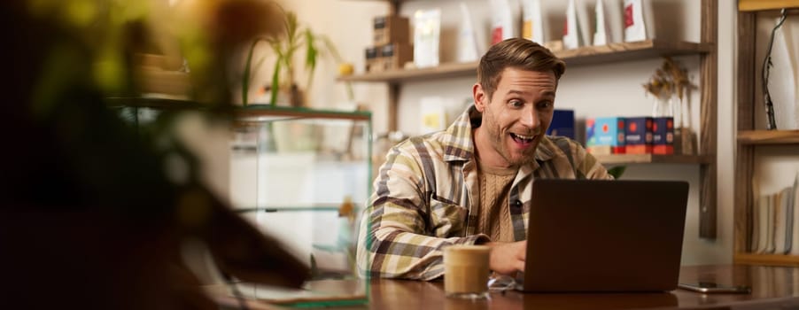Lifestyle portrait of young businessman, freelancer sitting in cafe, drinking coffee and using laptop, working on online project, digital nomad doing his task, has remote job, looking satisfied.