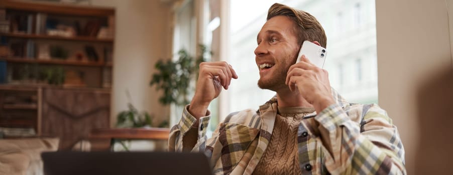 Portrait of young businessman, freelancer working remotely from coffee shop in city, sitting in chair with laptop, answer a phone call, talking on mobile to someone.