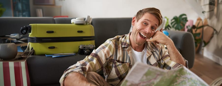 Portrait of handsome smiling young man, tourist shows call me gesture, phone hand, holding travel map, inviting to go on holiday, packed suitcase.