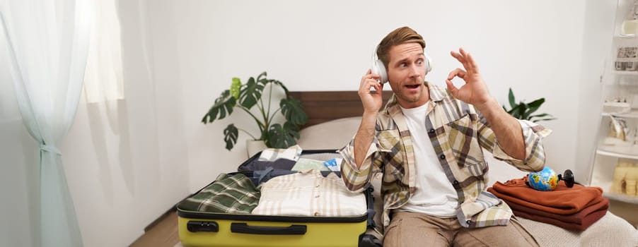 Portrait of handsome blond man, tourist goes on holiday, listens music in wireless headphones, packs clothes in suitcase, chooses vacation destination, shows okay, good hand gesture.