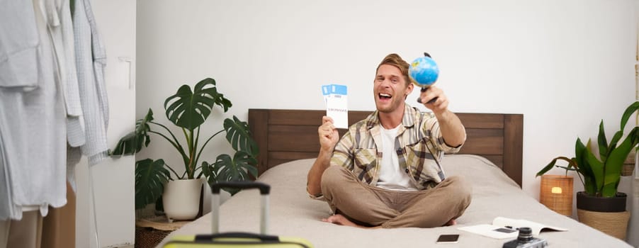 Excited, handsome young man sits on bed, shows globe and tickets, has suitcase, camera packed for holiday, goes on vacation, happy to go on holiday. Tourism concept