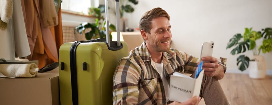 Portrait of handsome smiling man with suitcase, looks at smartphone, holds travel tickets and passport. Tourism and travelling concept