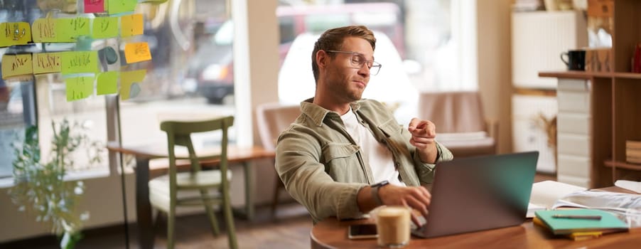 Portrait of handsome man in glasses, sits in cafe, points finger at laptop screen, video chats, connects to online meeting, feeling confident, working remotely.