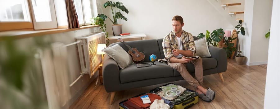 Portrait of young man packing for a trip, going on vacation, putting clothes inside suitcase, preparing for holiday.