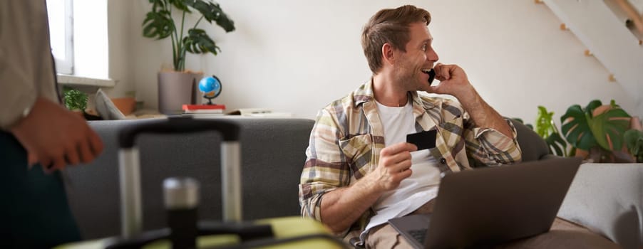 Image of guy calling someone, holding credit card and using laptop, has suitcase and camera packed for holiday. Tourism concept