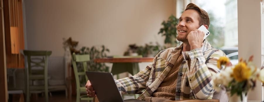 Image of young professional, cafe visitor working, sitting with laptop and talking on mobile phone to client. A guy making an enquiry buying something online, calling a friend.