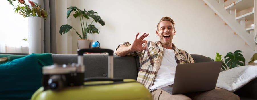 Happy man booking holiday online on laptop, paying for vacation on travel agency website, buying flight tickets in internet, sitting in living room with suitcase.