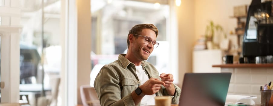 Portrait of handsome, smiling young man, online tutor, businessman working in cafe remotely, waving hand at laptop, connects to video chat or meeting.
