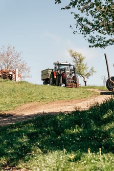 A tractor is seen maneuvering on a farm, towing a cargo trailer along a winding dirt path. The surrounding area features lush green fields and scattered trees, creating a serene rural atmosphere.