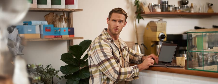 Portrait of man working in cafe, looking confused at camera, sitting with laptop and glass of coffee, staring puzzled at you.