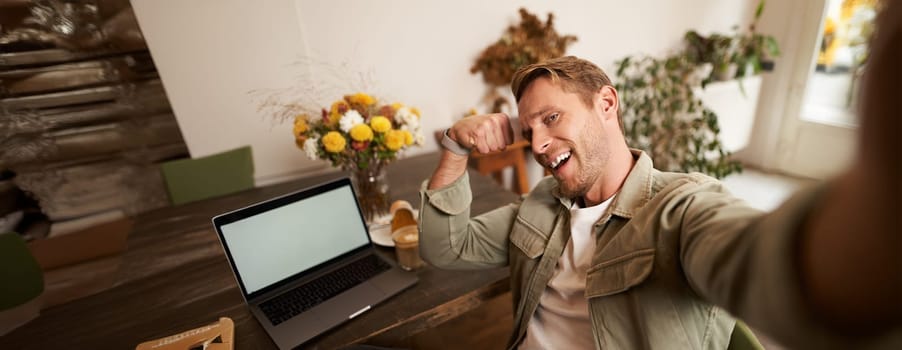 Portrait of handsome and confident man taking a selfie, flexing biceps, shows-off his muscles, sits in cafe in front of laptop, smiling at camera.