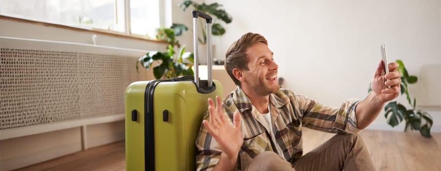 Portrait of happy tourist, man takes selfies before his flight, photos with suitcase, uses smartphone to post pictures on social media.