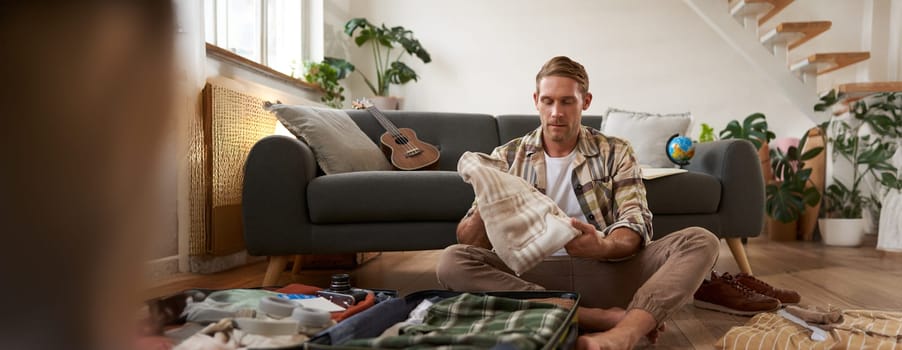 Travelling and tourism concept. Happy young man, sits on floor with luggage opened, packing suitcase for holiday, going on vacation, putting clothes inside a bag.