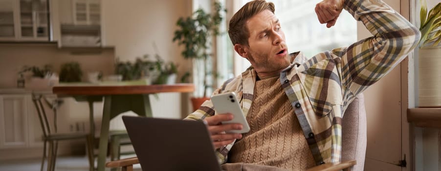 Portrait of young athletic guy sitting in cafe with laptop and smartphone, showing his biceps, flexing muscles and looking pleased at himself.