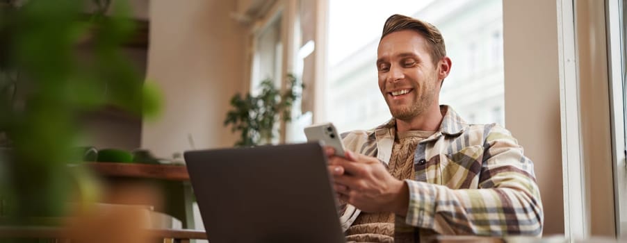 Close up portrait of handsome smiling man with laptop, cafe visitor sitting in chair, using smartphone, looking happy, messaging with friends while enjoying cup of coffee.
