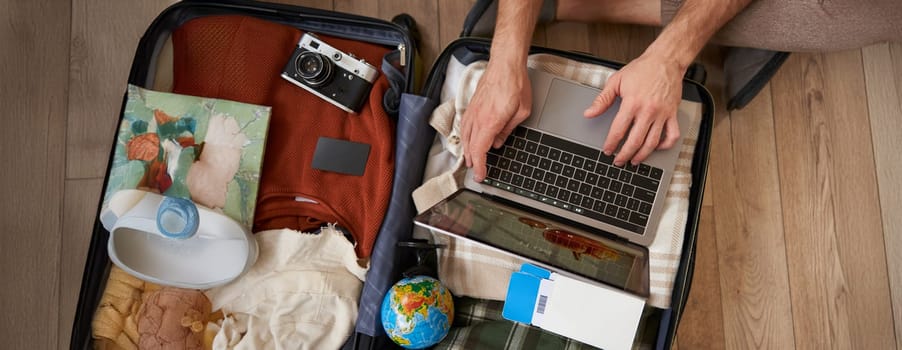 Close up portrait of male hands using laptop on top of opened suitcase, man packing for holiday, tourist with globe, booking vacation on travel agency website, picking a tour. Tourism concept