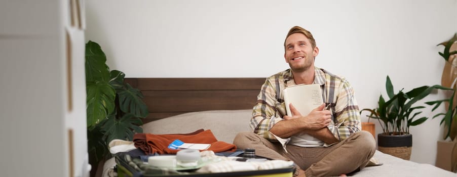 Portrait of thoughtful young man, tourist planning his vacation, sitting with open suitcase on bed, making notes, writing to do check list in notebook.