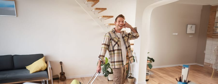 Full length photo of happy guy with suitcase, walking in apartment, shows phone call gesture. Copy space