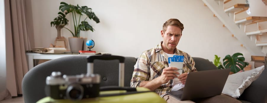 Portrait of frowning man, looking concerned at his flight tickets and passport, has laptop opened on his laps.