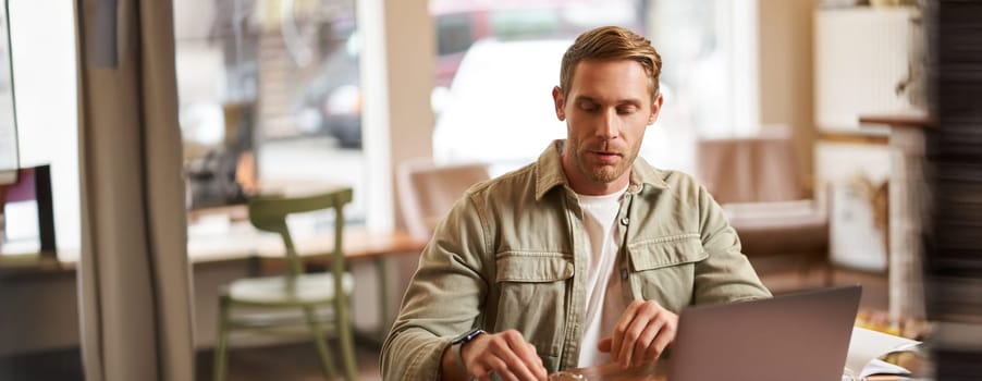 Image of young man, concentrated on his task, sitting with laptop in cafe, putting on glasses.