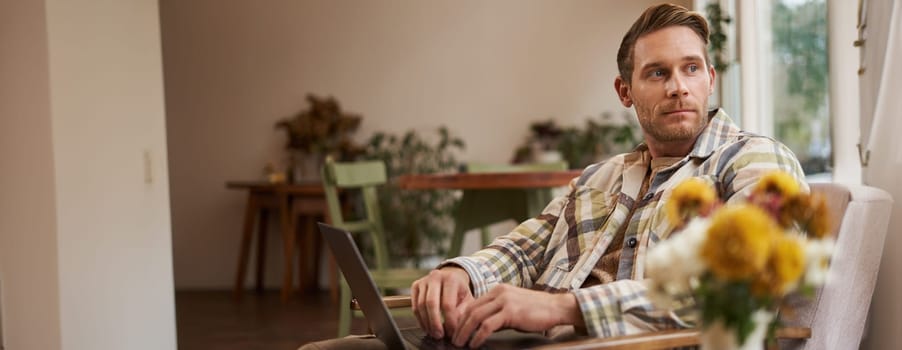 Portrait of handsome young man, working in cafe, sitting with laptop and coffee in hand, has remote job and using local co-working space instead of going to the office.