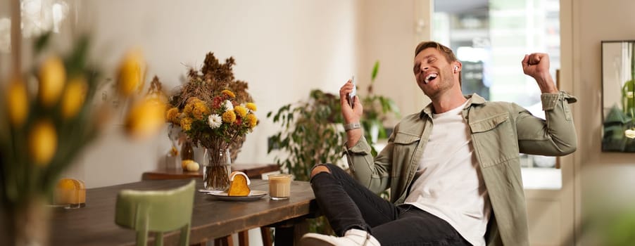Portrait of handsome happy man in cafe, listens to music in wireless earphones, holding smartphone, connects to public wifi and enjoys favourite song, relaxing in coffee shop.